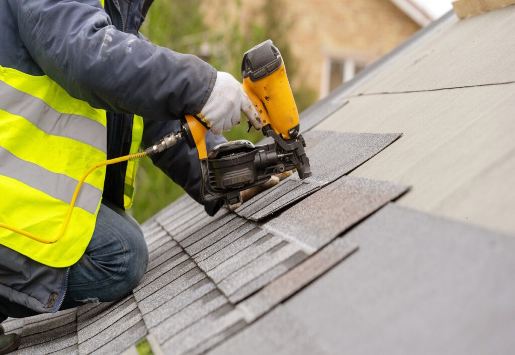 A roof worker in special protective work wear and gloves, using air or pneumatic nail gun and installing shingles on top a roof of a new house under construction, CK Contracting, New Custom Home Build, Mount Pleasant, SC.